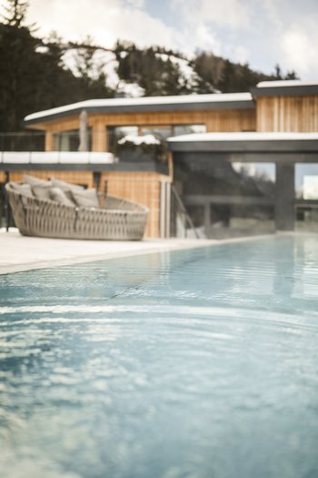 Il vostro hotel con spa tra le Dolomiti dell’Alto Adige Piscina esterna con casa in legno moderna e poltrona in vimini imbottita sullo sfondo
