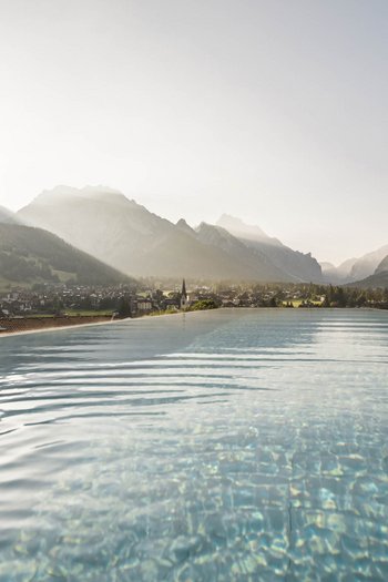 Wellnesshotel in Südtirol: Auf in den Traumurlaub! Infinity-Pool mit Blick auf Berge und Dorf bei Sonnenuntergang