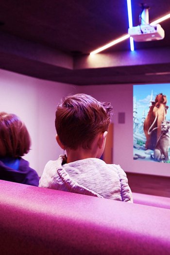 Familienurlaub in den Dolomiten – Natur, Erholung & Abenteuer Kinder schauen einen Zeichentrickfilm im Heimkino auf einer rosa Couch
