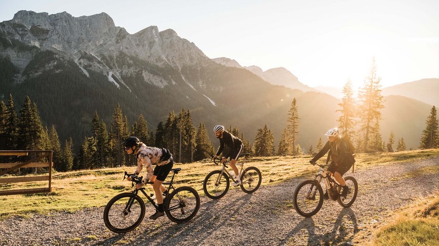 Bici gravel a San Vigilio Tre ciclisti su un sentiero di montagna al tramonto con montagne sullo sfondo