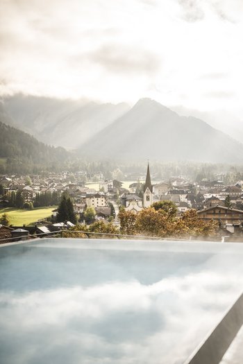Hotel con infinity pool in Alto Adige: l’Excelsior Vista di un paese alpino con chiesa e montagne nebbiose da una piscina