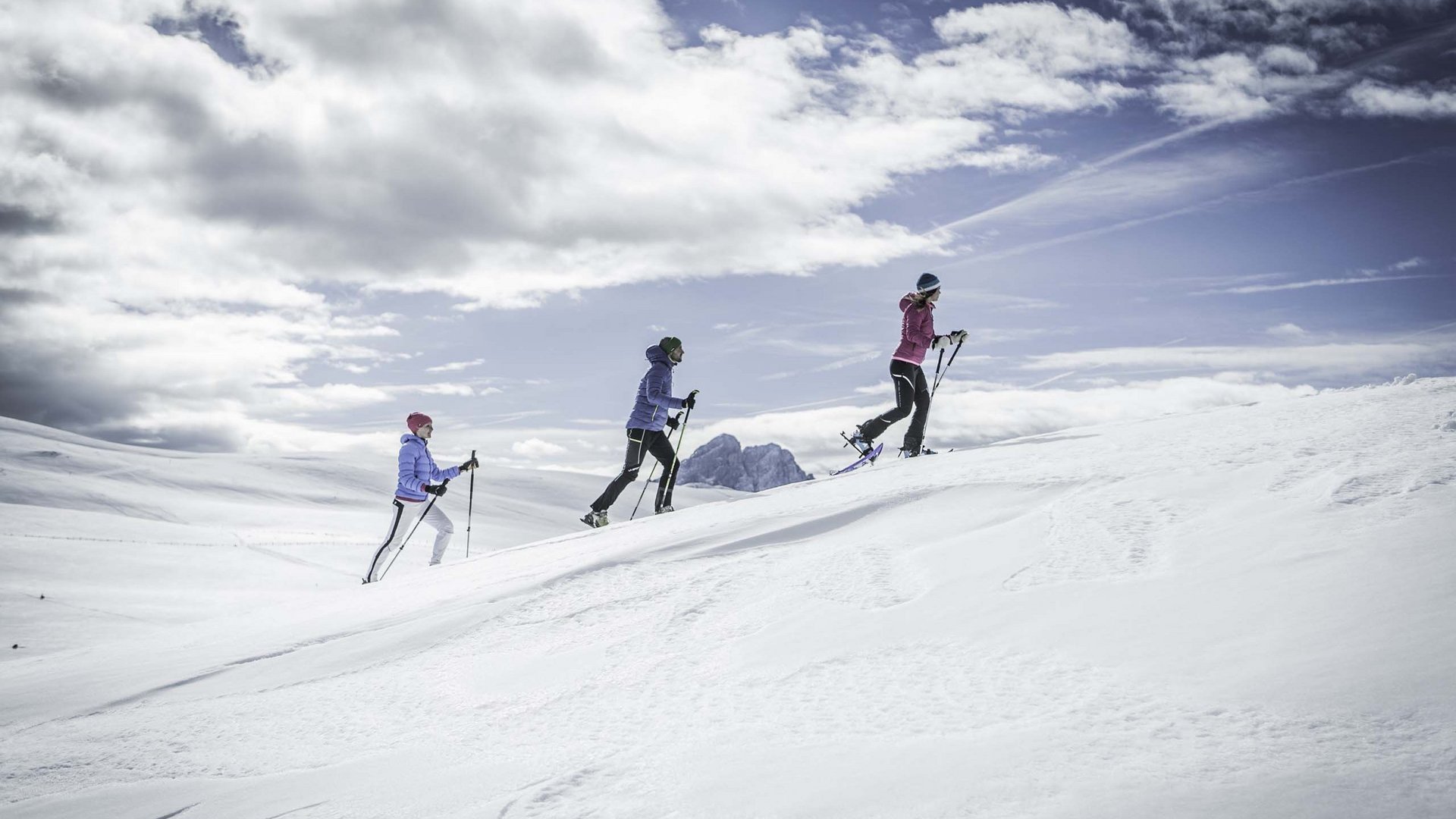 Kronplatz, Südtirol – Aktivitäten & Highlights Drei Personen beim Schneeschuhwandern auf verschneitem Hügel unter bewölktem Himmel