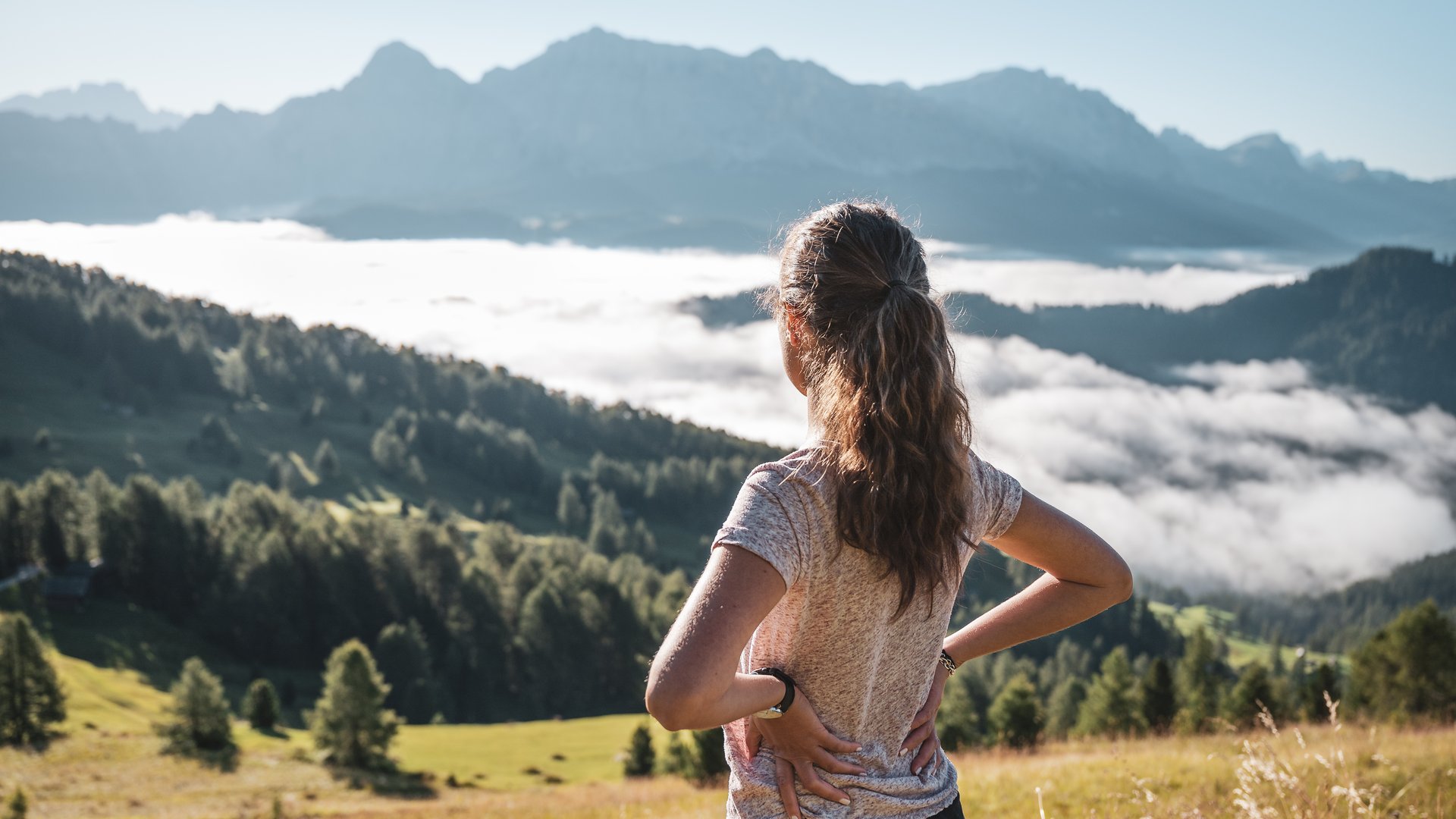 Excelsior Dolomites Life Resort: Ihr Sporthotel in den Dolomiten Frau steht in den Bergen und blickt auf neblige Täler und Berggipfel